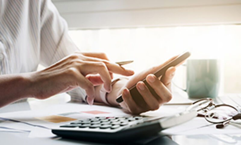 "Eine Frau im Büro, die mit einem Stift auf einem Smartphone arbeitet. Vor ihr liegen ein Laptop und finanzbezogene Dokumente – symbolisch für Preisberechnung."