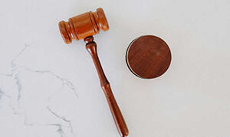 "Ein brauner Richterhammer mit dünnem Griff und breitem Kopf, liegend auf einer weißen Oberfläche. Neben dem Hammer liegt ein Holzblock, auf den der Hammer geschlagen wird – symbolisch für Gesetze."