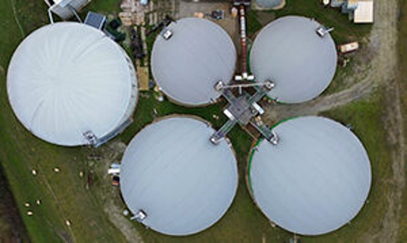 "Eine Luftaufnahme einer Biogasanlage mit mehreren großen weißen Rundbehältern auf einem grünen Feld. Die Anlage dient der Erzeugung erneuerbarer Energie aus Biomasse."
